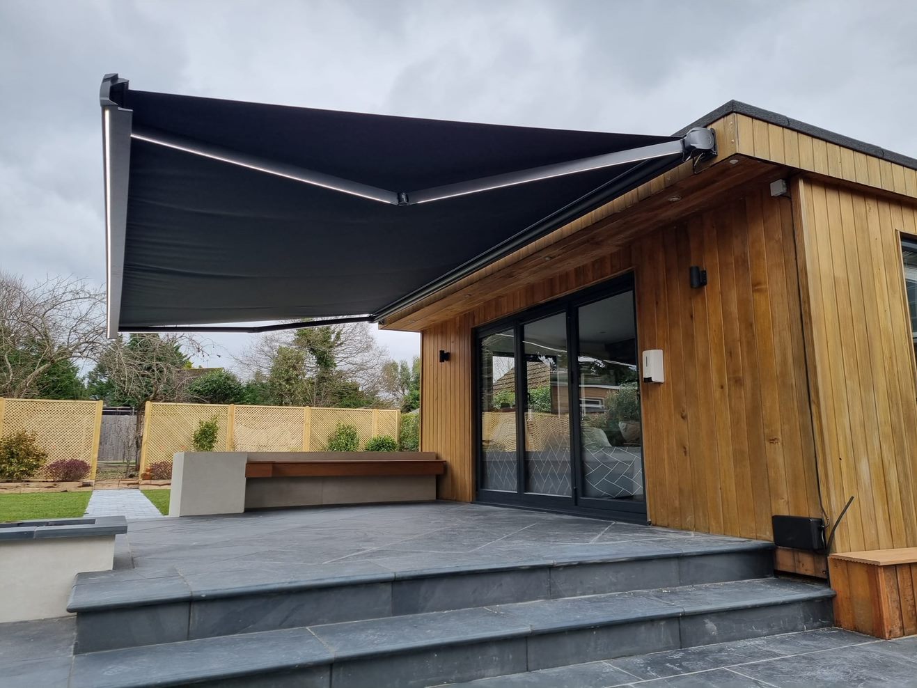 Wide grey compact cassette awning spanning the whole rear elevation of a modern London extension above a garden dining table